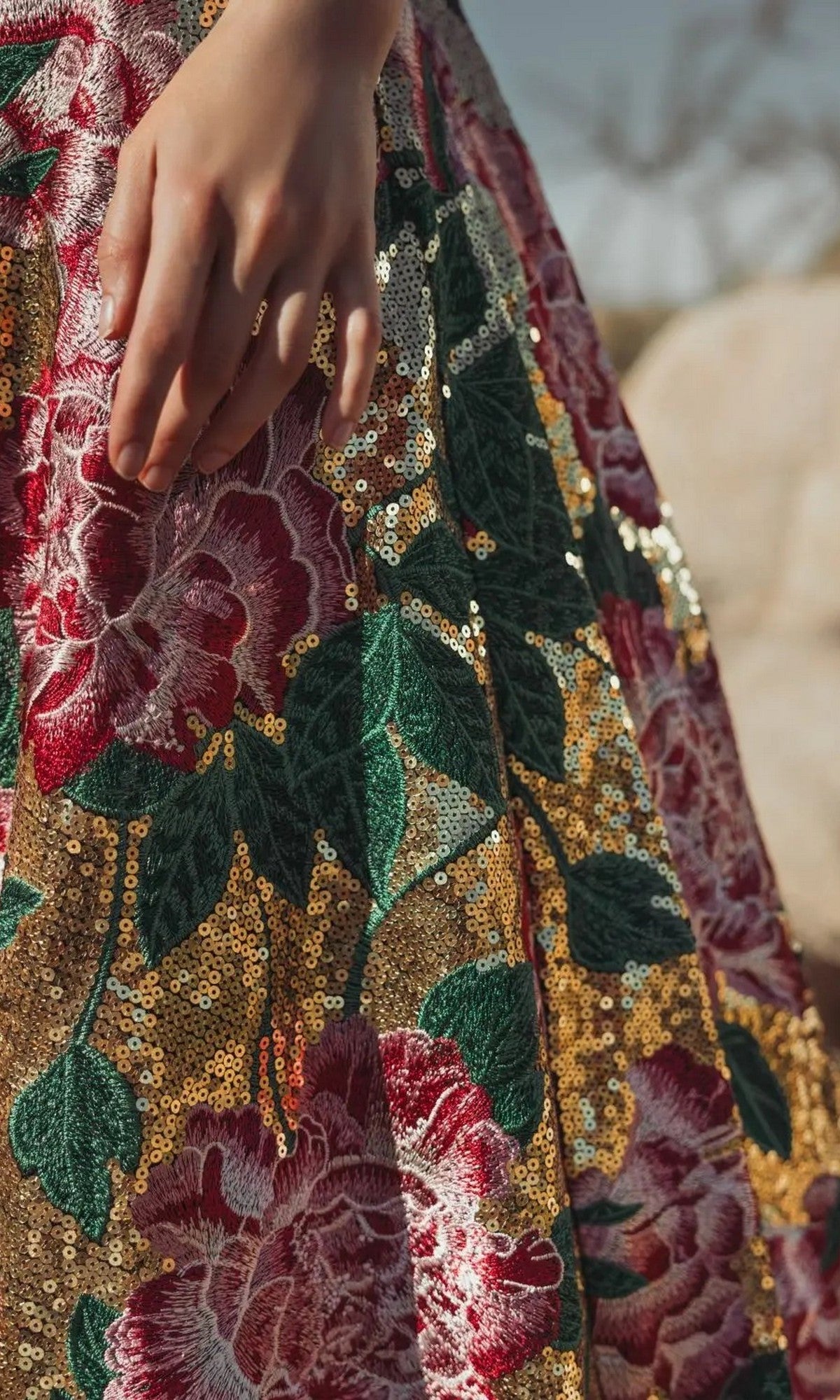 A close-up of a hand resting on the Mac Duggal 21194 long formal dress, featuring gold sequins and bold floral embroidery in red, green, and pink.